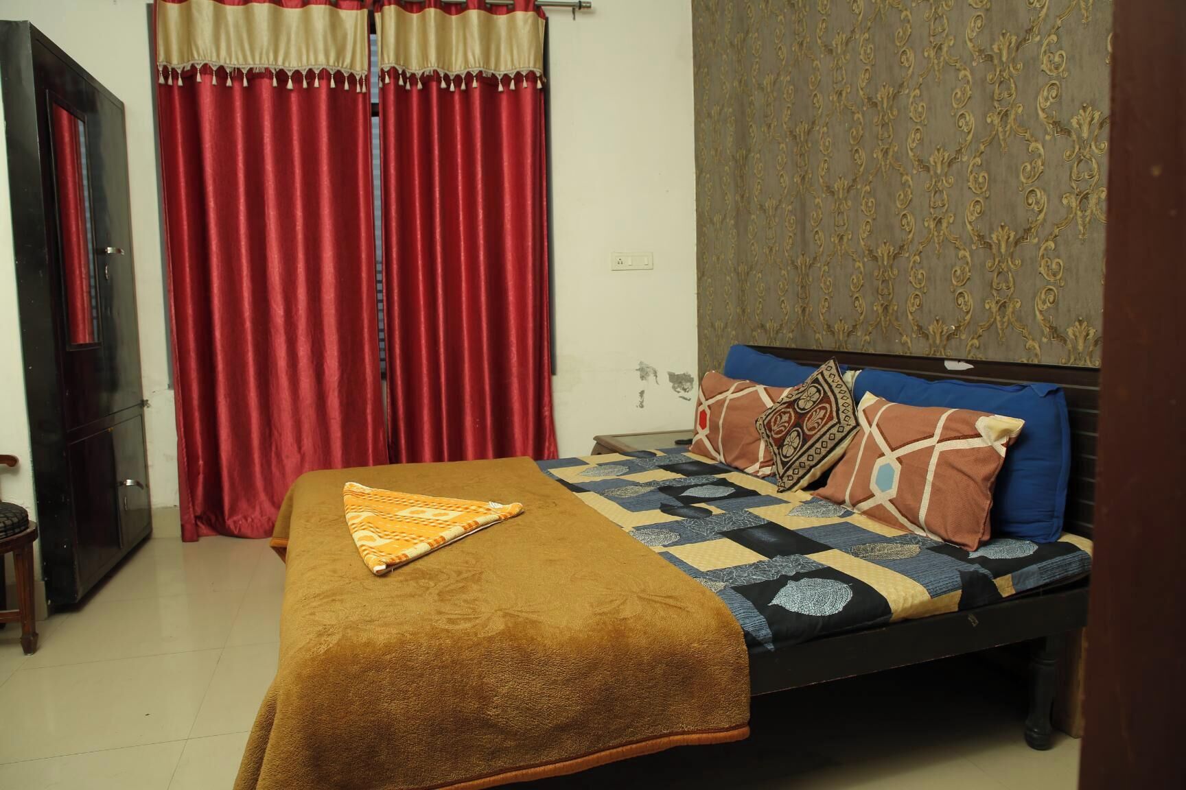 A bedroom with a bed, red curtains, a patterned wall, and a brown throw blanket.