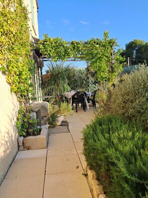 Un sentier mène à un espace de repos abrité, surmonté d’une pergola, et entouré de diverses plantes et arbustes.