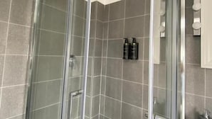 A glass-enclosed shower with a white towel hanging on the door, grey tiled walls, and a small shelf with toiletries. - Bari LandLord 242 (Bari)