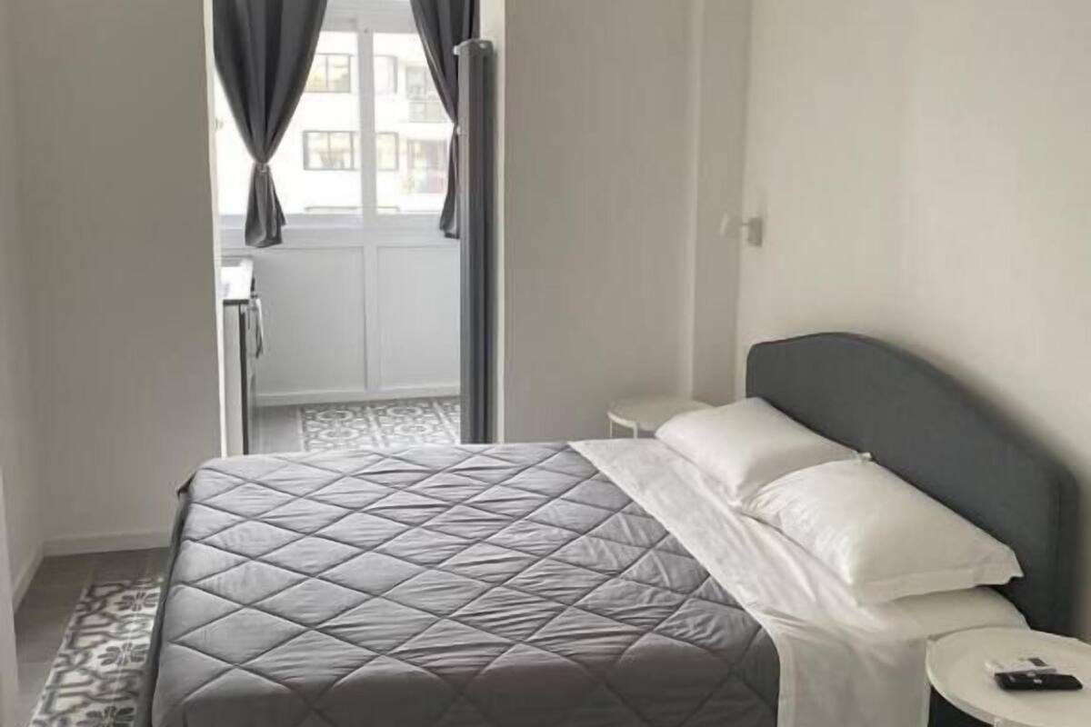 A hotel room with a bed, a small bedside table, a window with curtains, and a patterned floor.