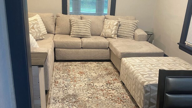 Ein gemütlicher Wohnraum mit Ecksofa, einem gemusterten Teppich, einem kleinen Tisch und einem Fenster mit Jalousien.