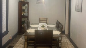 Ein Esszimmer mit einem rechteckigen Tisch, gedeckt für vier Personen, umgeben von Stühlen. Über dem Tisch ist ein Deckenventilator zu sehen, und links befindet sich ein Bücherregal mit Dekorationen.