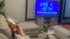 Ein Wohnzimmer mit Fernseher, der eine Nachrichtensendung zeigt, Deckenventilator und Fenster mit Jalousien.
