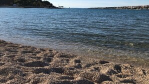 A beach with clear water, pebbly shore, and a distant shoreline with buildings. - Apartment Oleander (Novalja)
