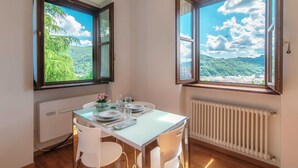 Uma área de refeições com uma mesa posta para dois, grandes janelas com vista para colinas verdes e um céu claro.