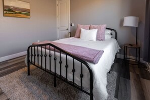 Una cama bien hecha con estructura metálica negra, ropa de cama blanca y una manta morada.