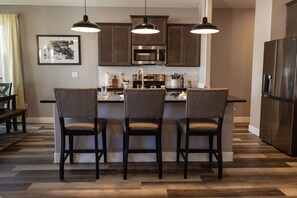 Una cocina moderna con una isla central, tres taburetes de bar, iluminación colgante y electrodomésticos de acero inoxidable.