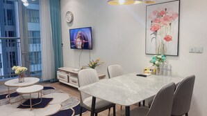 A modern living room with a dining area, a TV mounted on the wall, and a large window with curtains.