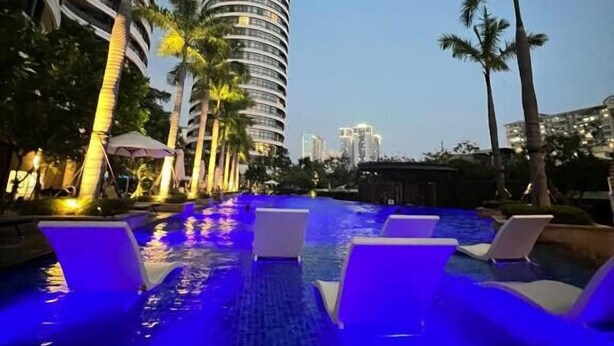 A swimming pool with water features and lounge chairs, surrounded by palm trees and modern buildings.