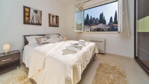 A bedroom with a bed, a nightstand, a window with a view of trees and buildings, and artwork on the wall. - 4B Apartments (Dubrovnik)