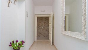 A narrow hallway with white walls, a tiled floor, a potted plant with purple flowers, and a large mirror with a textured frame. - 4B Apartments (Dubrovnik)