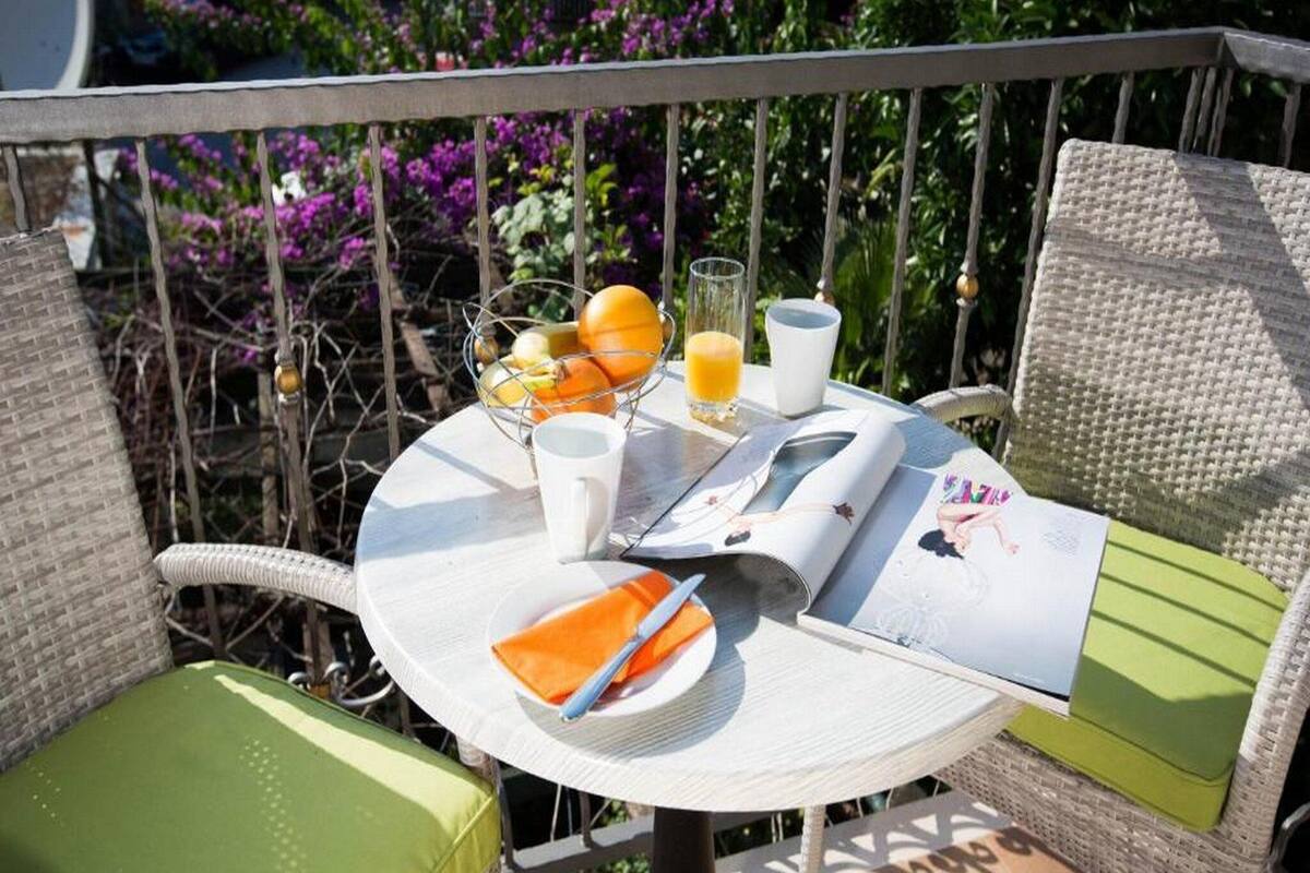 Une table ronde d’extérieur, sur laquelle on trouve un magazine, un panier de fruits, un verre de jus d’orange et une tasse blanche.
