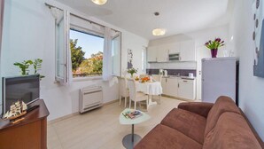 A modern living room with a brown sofa, a TV on a wooden cabinet, a dining area with a white table and chairs, and a kitchen in the background. - 4B Apartments (Dubrovnik)