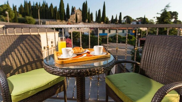 A balcony with wicker furniture, a small round table with breakfast items, and a view of trees and buildings. - 4B Apartments (Dubrovnik)
