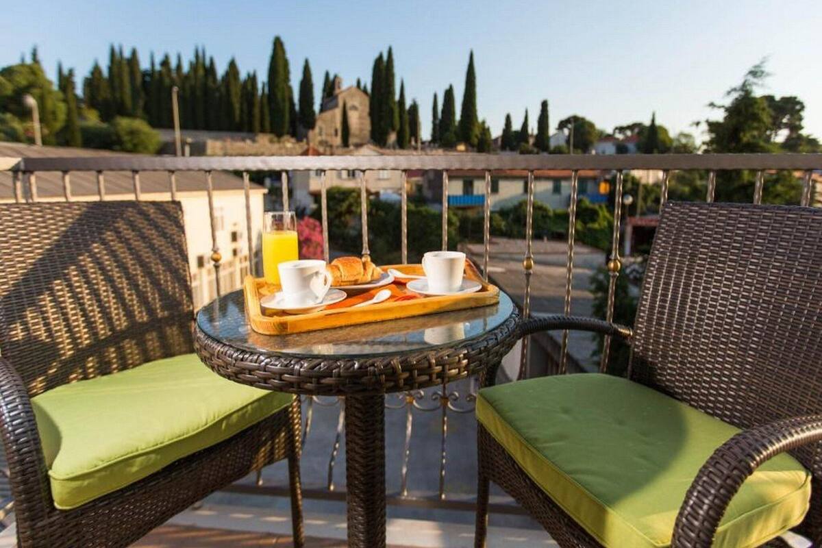 Un balcon agrémenté de meubles en osier, une petite table ronde avec des éléments pour le petit-déjeuner, et une vue sur les arbres et les bâtiments.