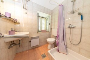 A bathroom with a sink, toilet, shower, and a window with blinds.
