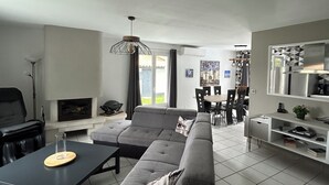 Ein modernes Wohnzimmer mit einem grauen Sofa, einem Couchtisch und einem Kamin. Im Hintergrund befindet sich ein Essbereich mit Tisch und Stühlen.