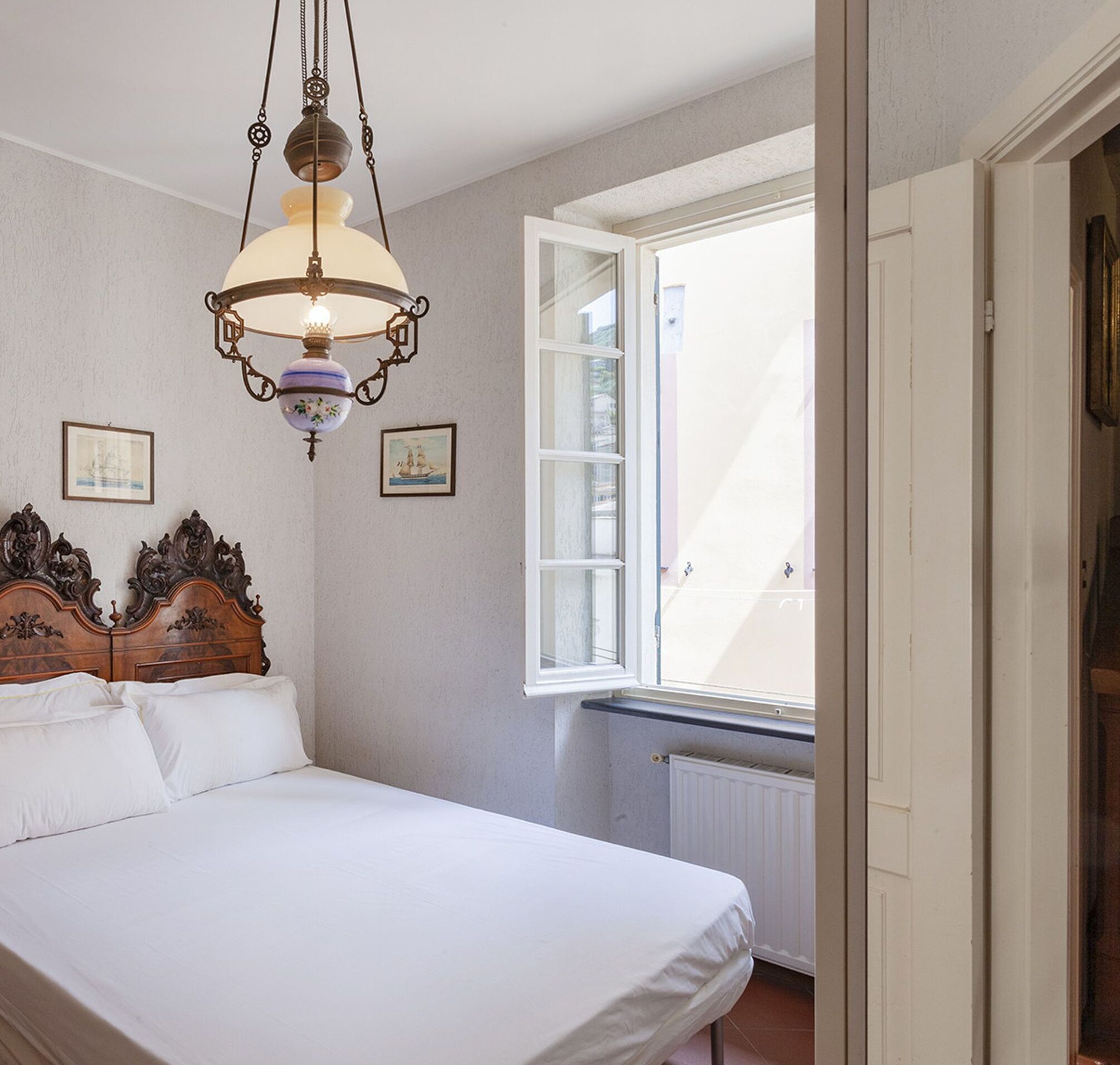 A bedroom with a large bed, a hanging light fixture, a window with a view, and two framed pictures on the wall.