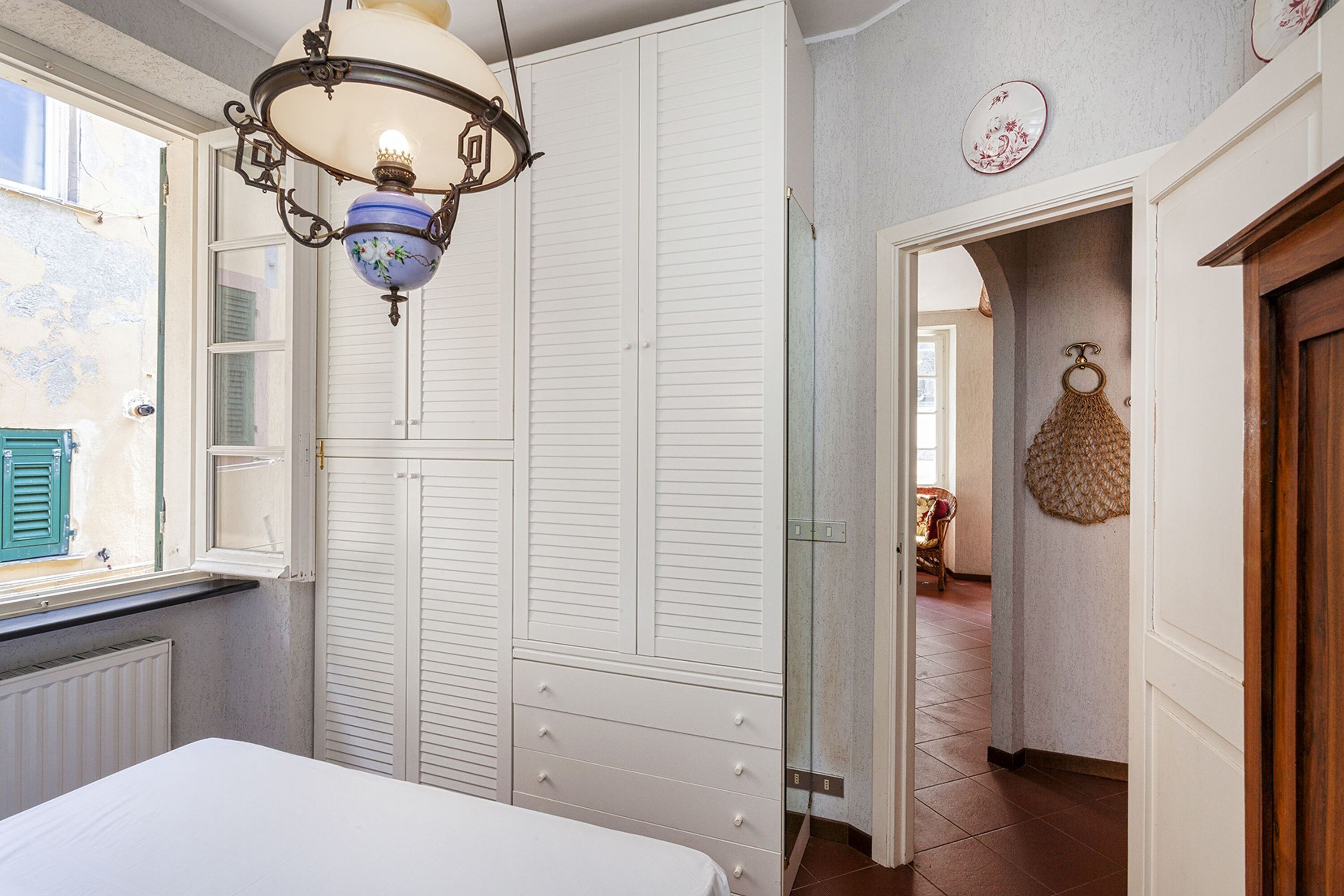 A bedroom with a bed, a wardrobe, a hanging light fixture, and a view through an open window.