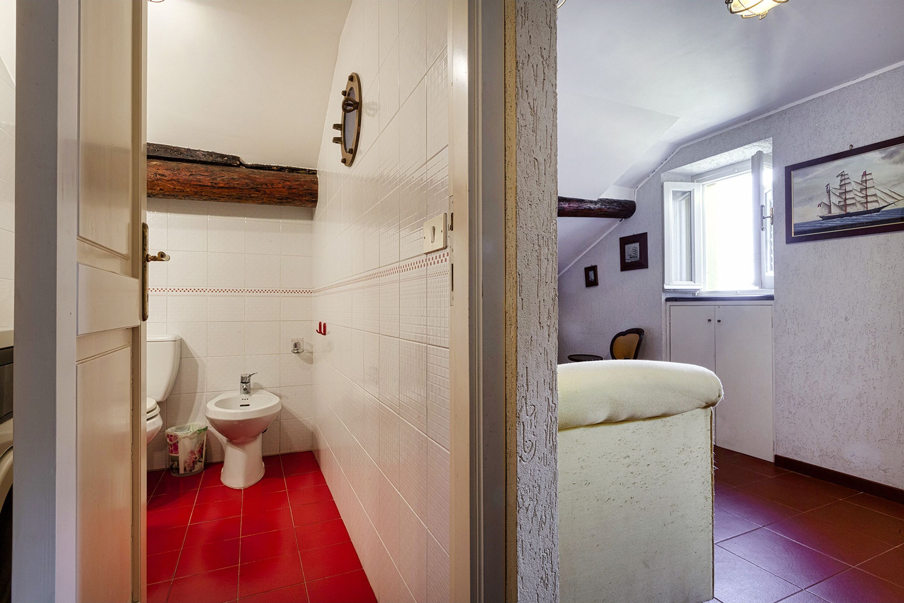 A bathroom with a red-tiled floor, white tiles on the walls, a white toilet, a bidet, and a window with a ship painting.