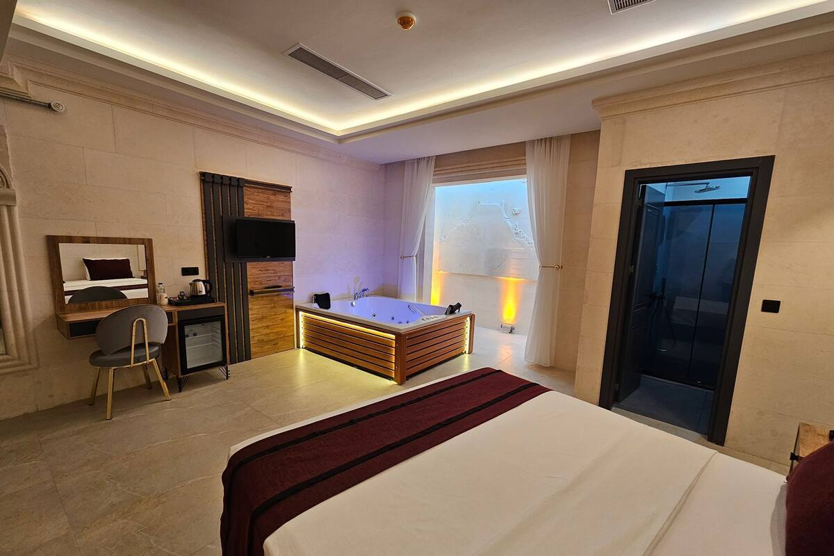 A hotel room with a large bed, a desk with a chair, a television, a bathtub, and a window with curtains.