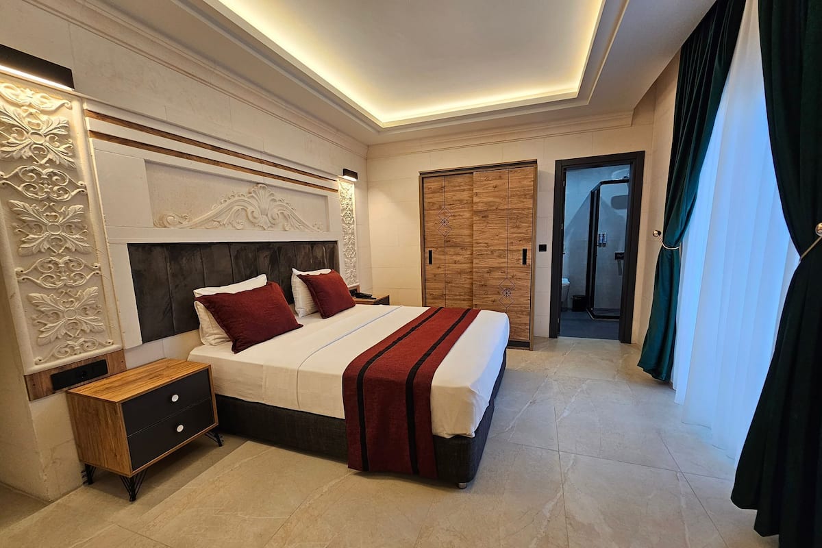 A modern hotel bedroom with a large bed, a nightstand, a wooden wardrobe, and a bathroom visible through an open door.