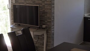 A modern living room with a flat-screen TV on a white stand, a stone-patterned wall, a dining table with chairs, and a small kitchen area with a refrigerator. - Apartments Profaca (Rab)