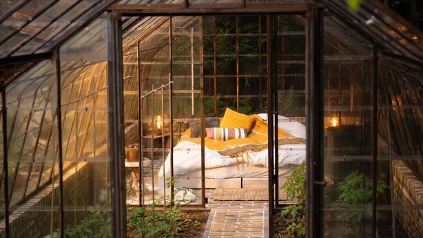 Une serre avec un parterre à l’intérieur, entourée de plantes et bordée d’un chemin en briques.