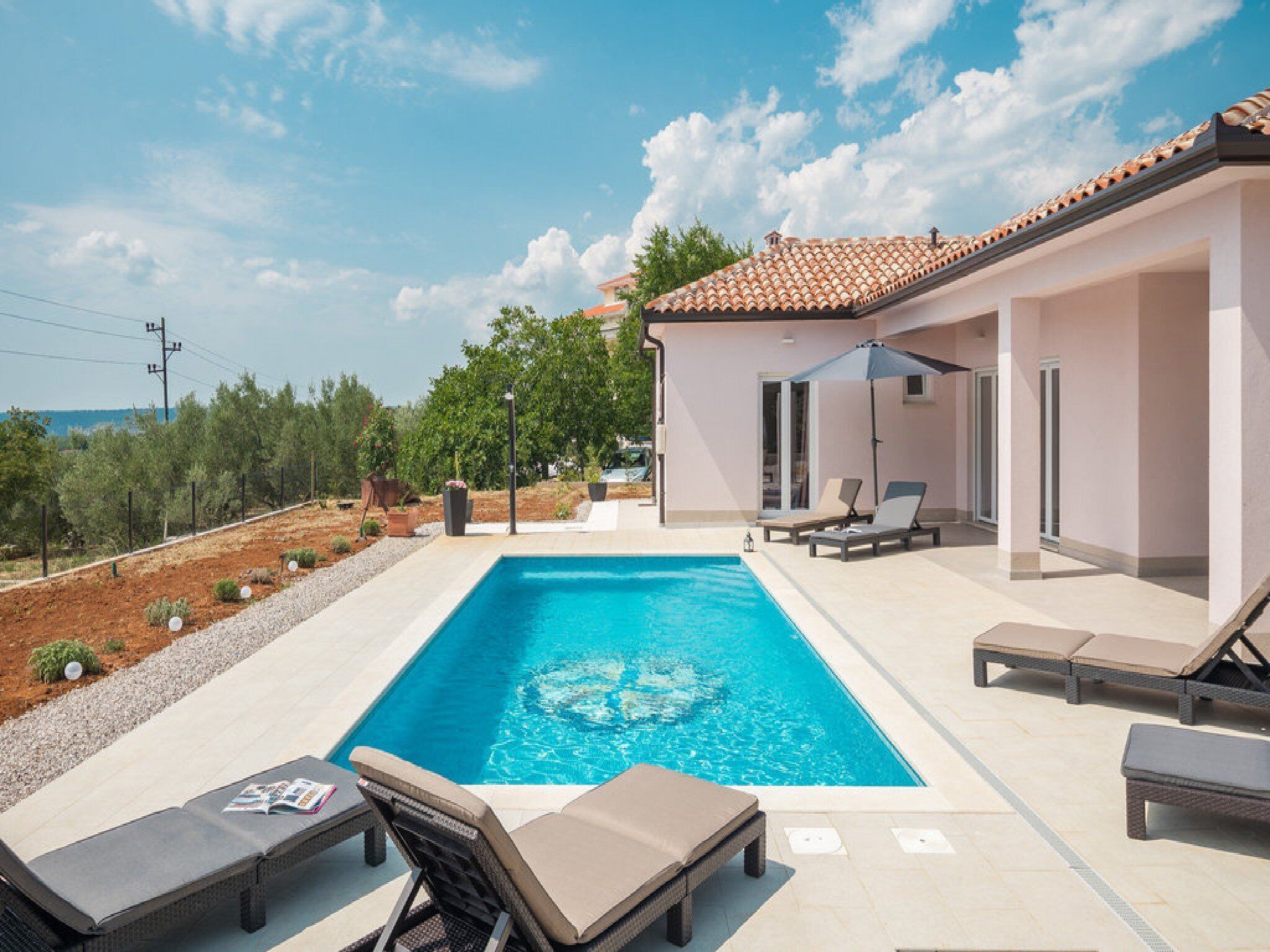 Piscina com espreguiçadeiras e área coberta, rodeada por um jardim e um edifício com telhado em azulejos.