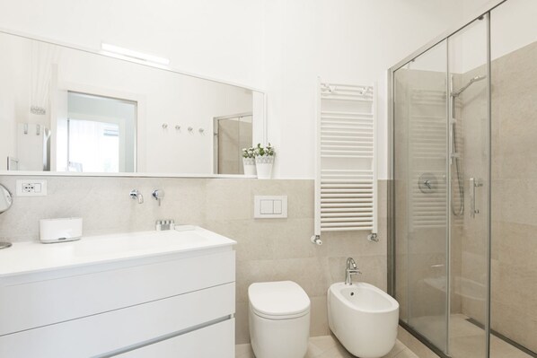 Un baño moderno con tocador blanco, inodoro, bidé, ducha con mampara de vidrio y un espejo grande.