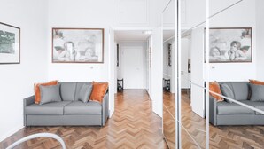 A modern living room with a grey sofa, orange cushions, and a herringbone wood floor. There are framed artworks on the walls and a large mirror reflecting the room. - Easylife - Residential apartment on Navigli area (Milano)