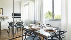 A modern dining area with a glass table set for four, surrounded by black chairs. A large window provides natural light and a view of greenery outside. - Easylife - Residential apartment on Navigli area (Milano)