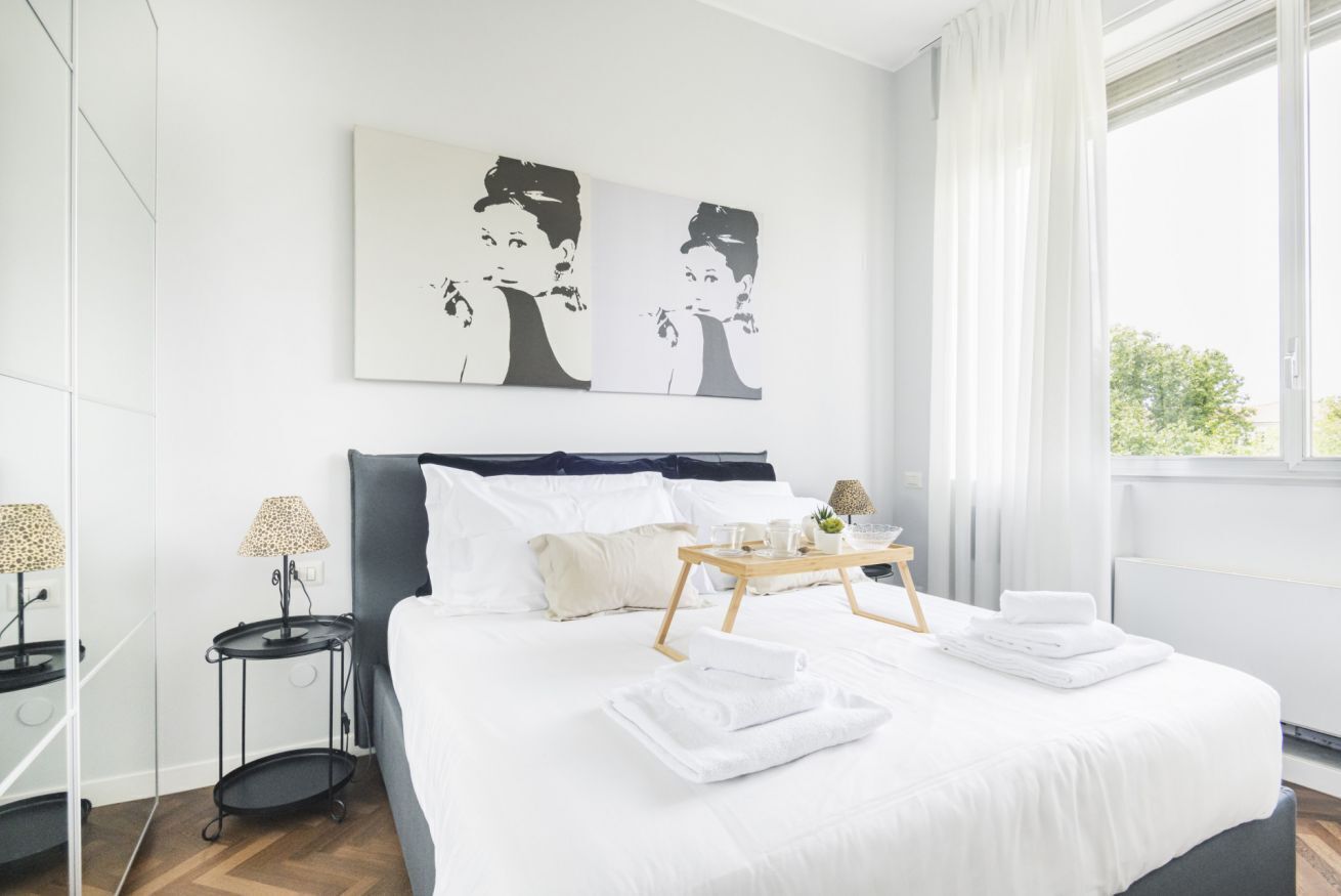 A modern hotel room with a large bed, a bedside table with a lamp, a tray with towels, and a window with curtains.