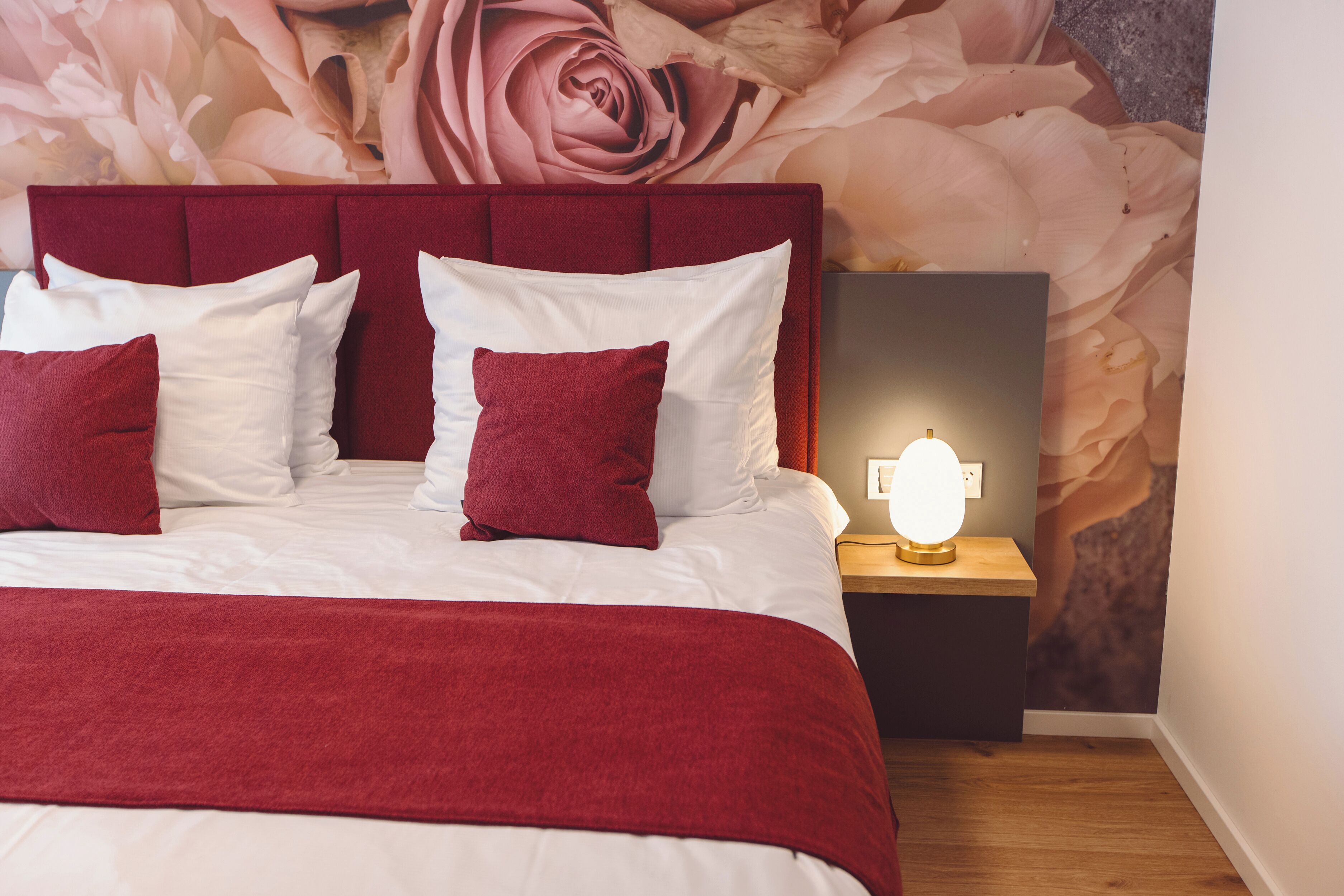 Un lit bien fait, avec une tête de lit rouge et du linge de lit blanc, une lampe de chevet et une fresque florale sur le mur.