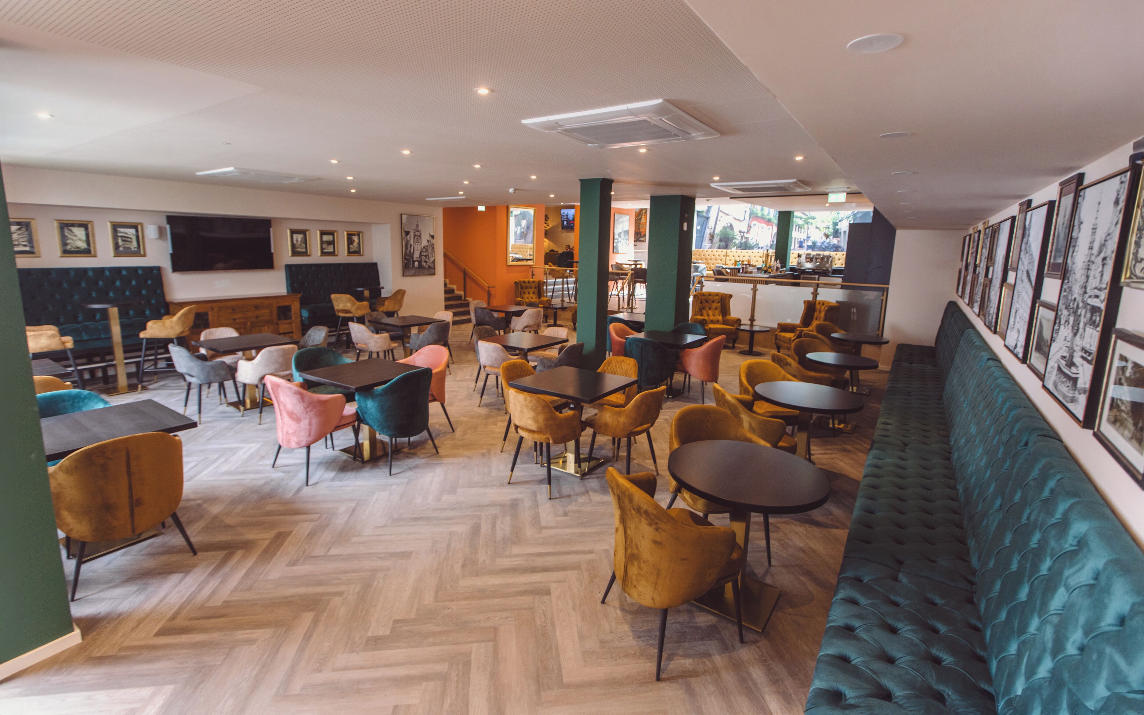 A modern cafe with wooden herringbone flooring, multiple seating areas including tables and chairs, and a large window providing natural light.