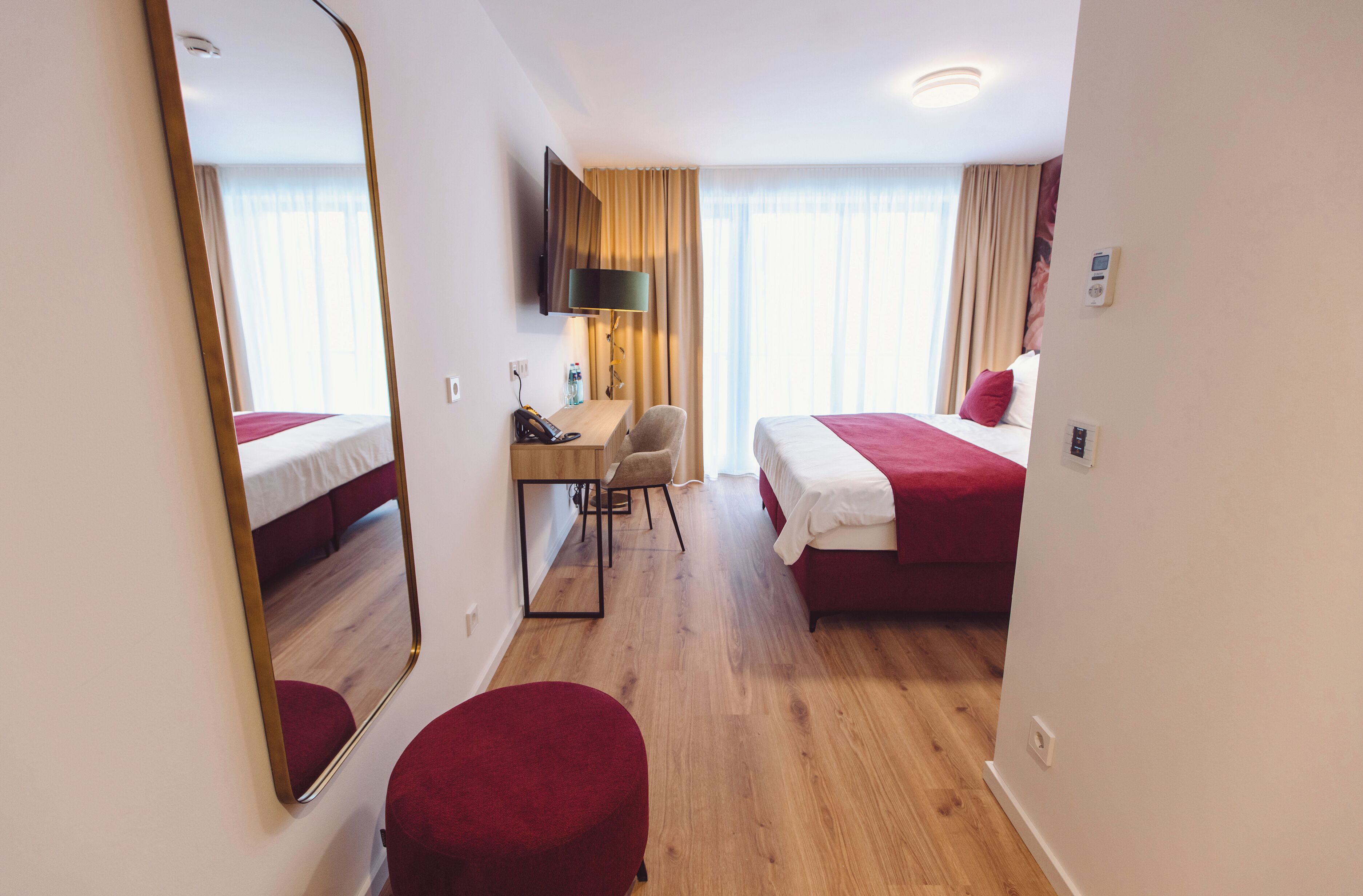 Una habitación de hotel con piso de madera, un escritorio con silla, una cama, un reposapiés rojo, un televisor montado en la pared y una ventana con cortinas.