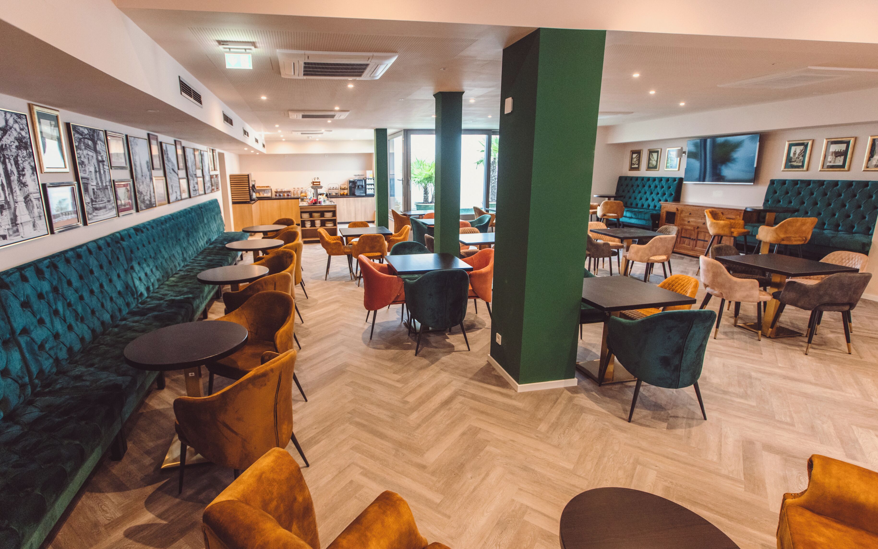 A modern cafe with a green cushioned bench, wooden tables, and colorful chairs.