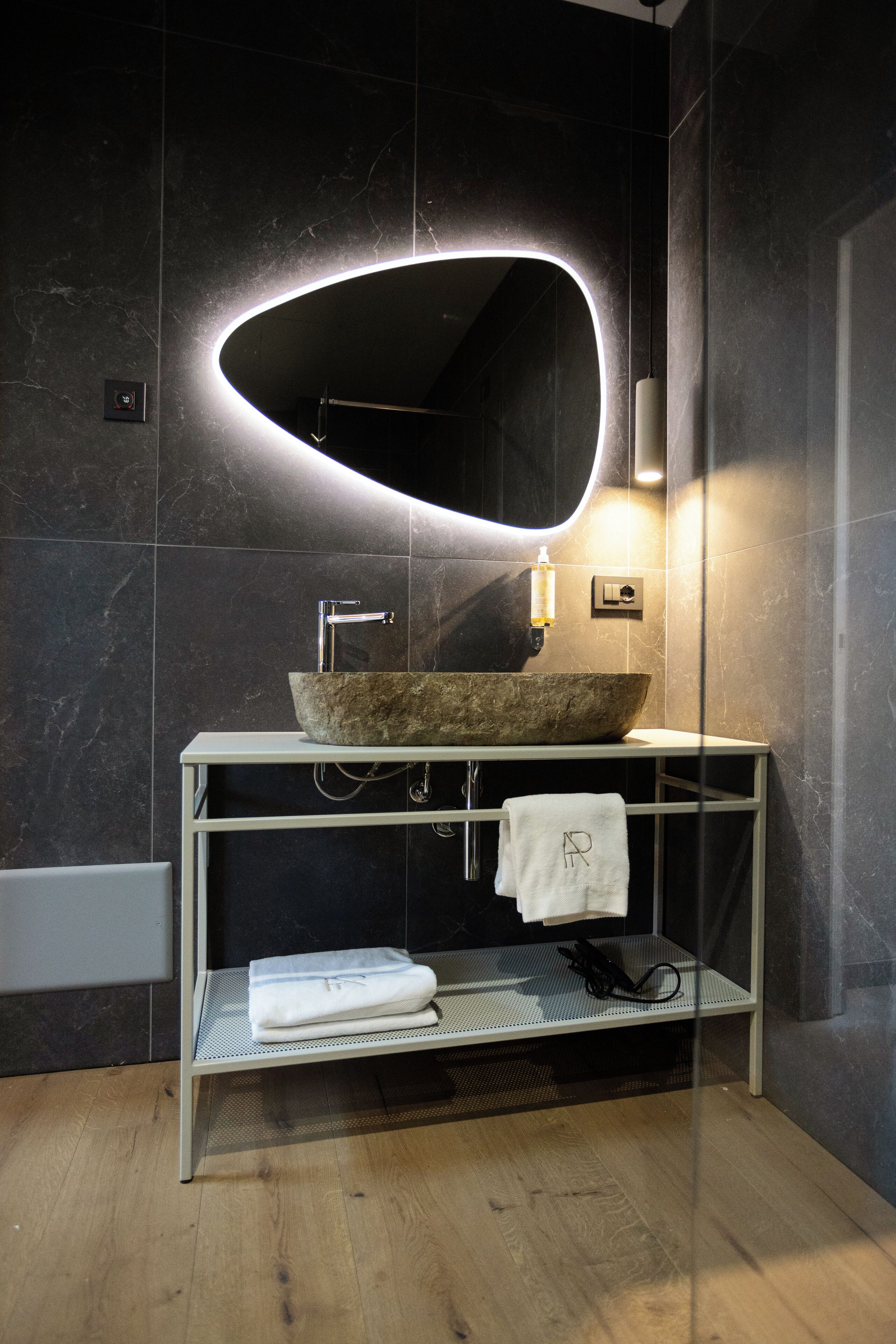 Un baño moderno con un espejo grande y ovalado, un lavabo de piedra y una estantería metálica con toallas.