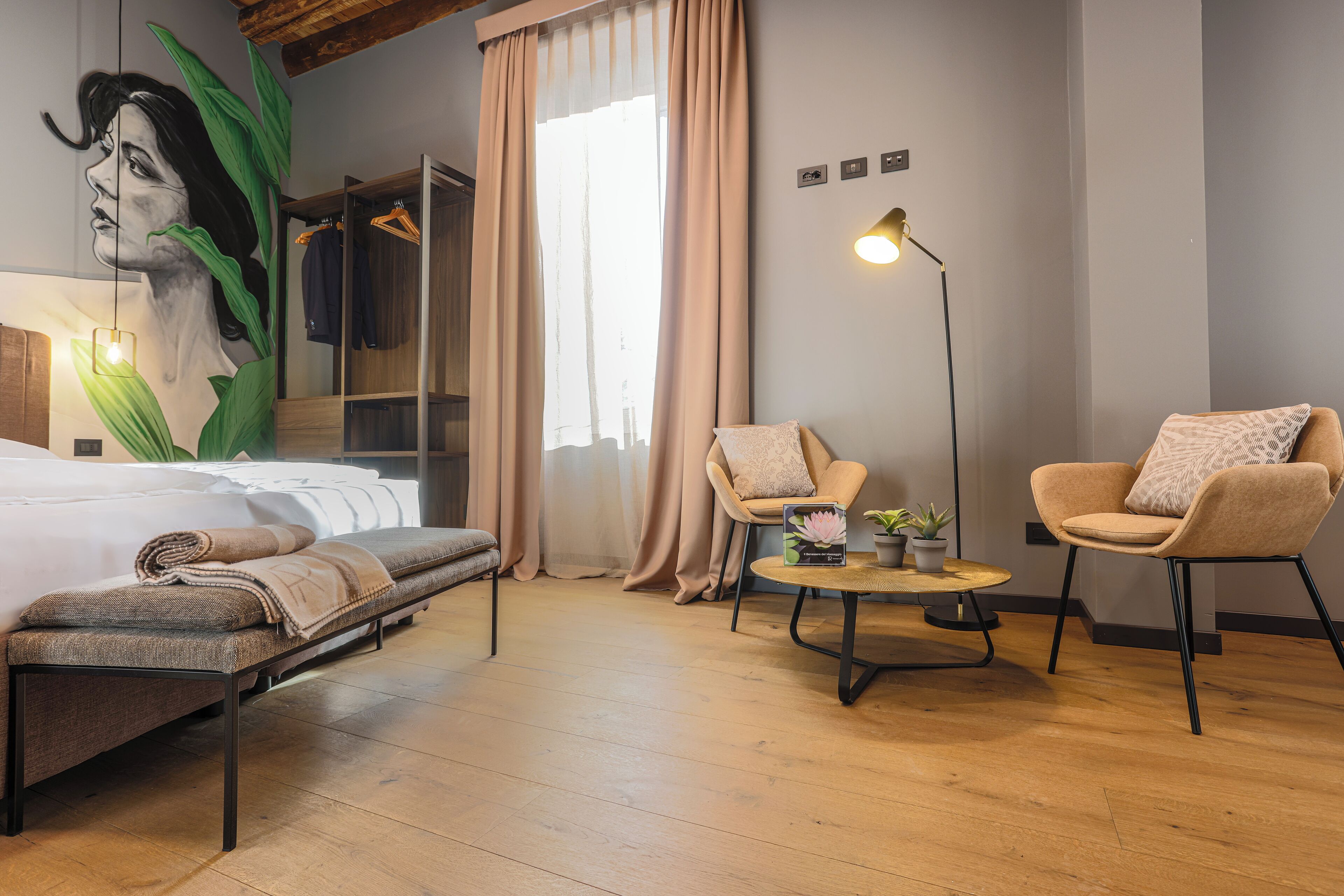 A modern hotel room with a bed, two armchairs, a small table with a plant, and a large window with curtains.