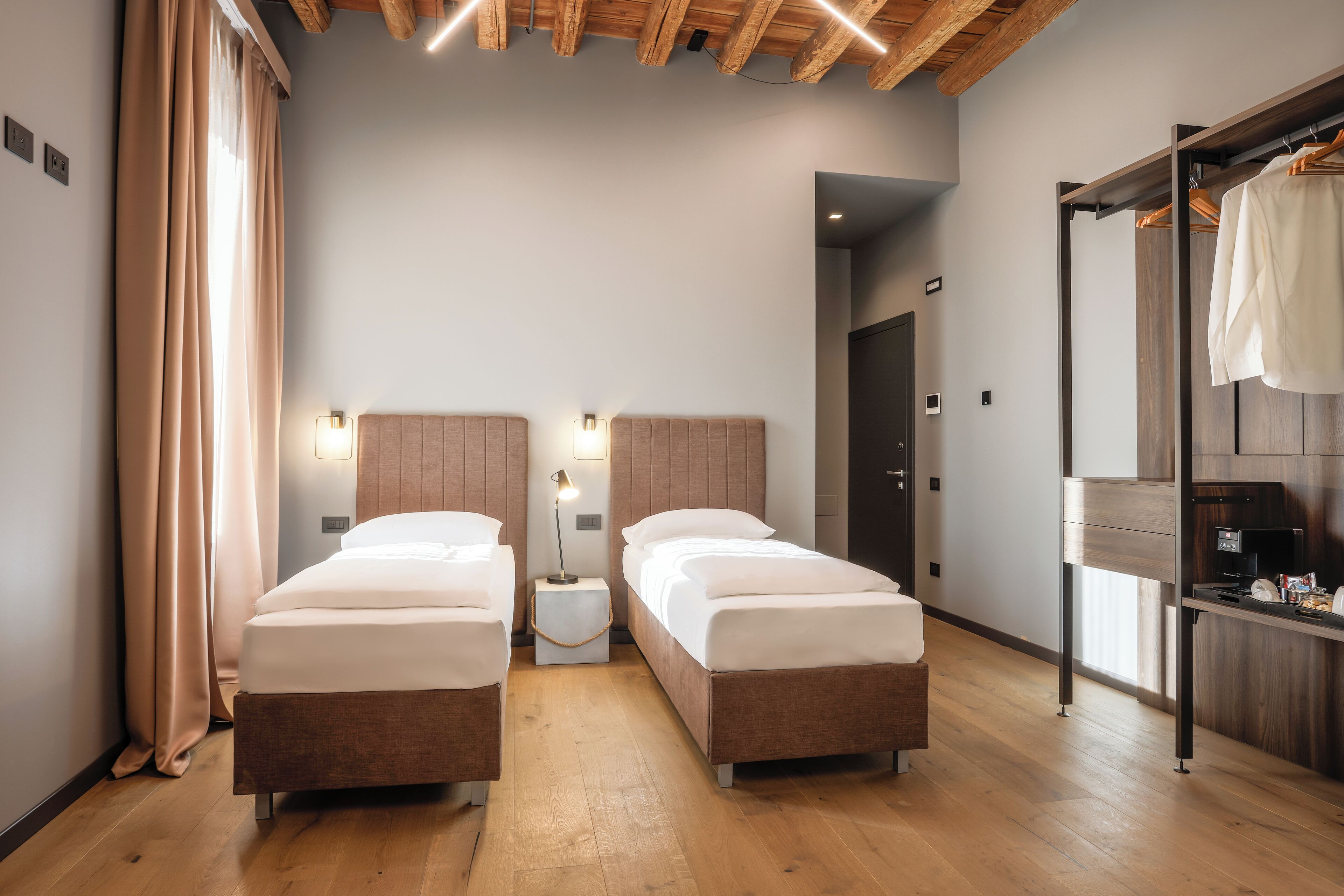 Habitación de hotel con dos camas, suelo de madera, un armario y una mesita de noche pequeña con una lámpara.