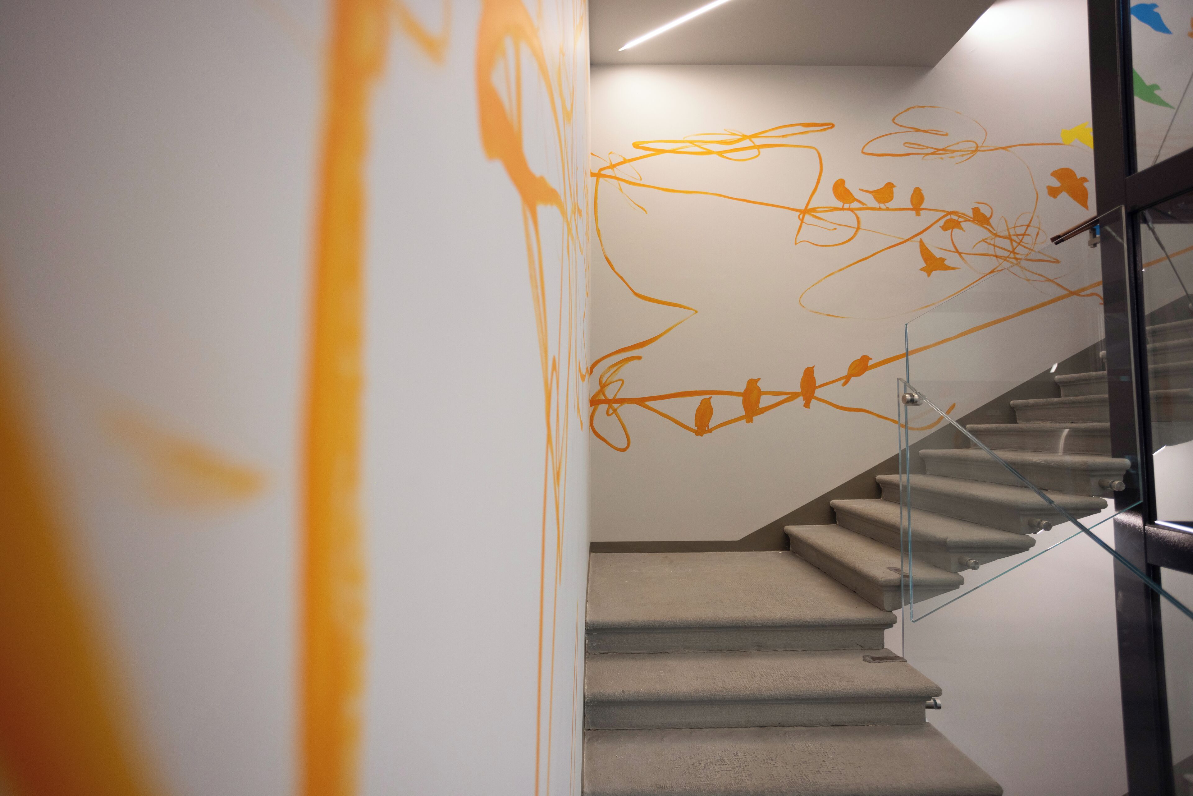 Una scala con ringhiera in vetro, un muro con graffiti arancioni astratti e un moderno apparecchio di illuminazione.