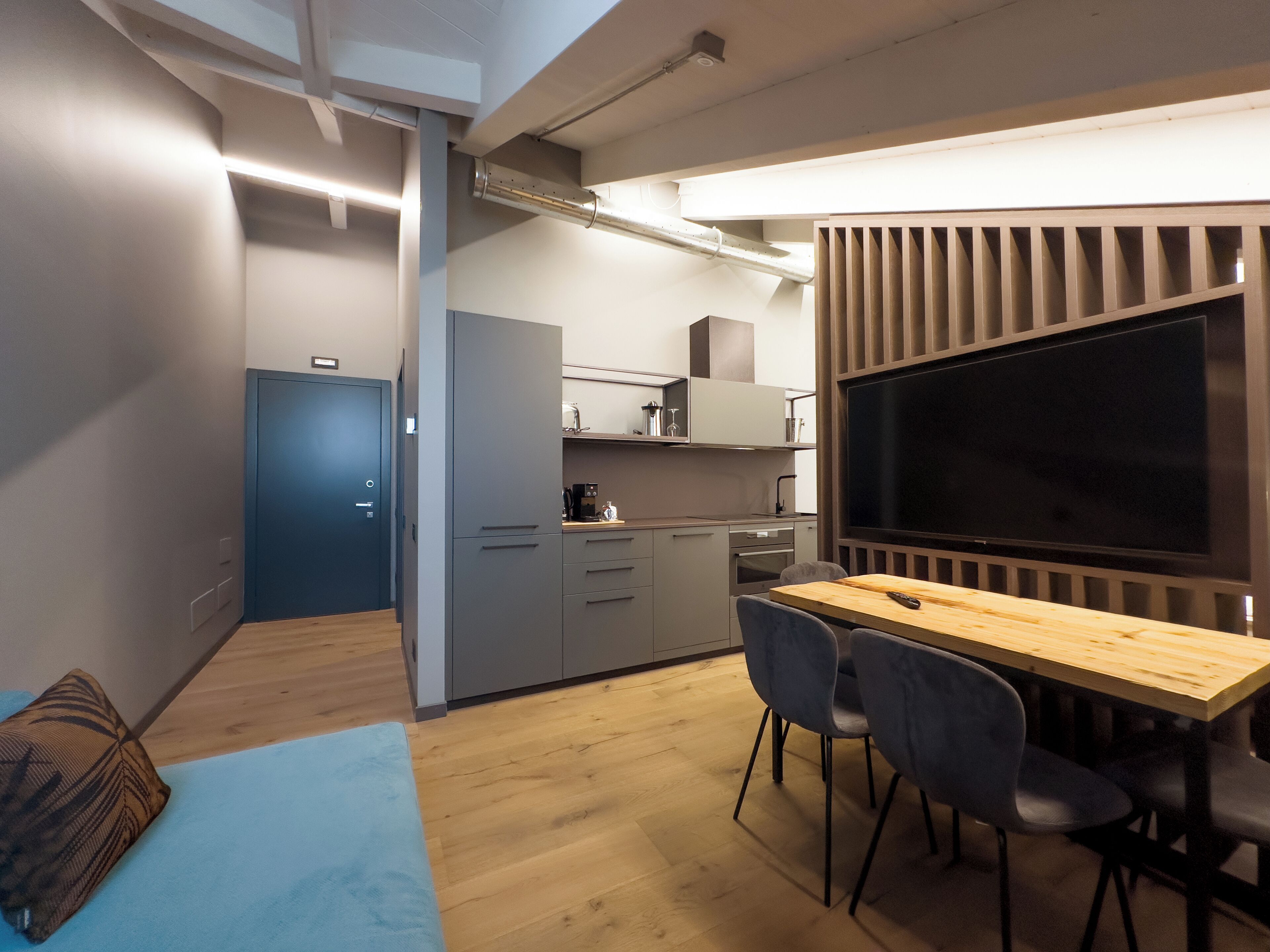 Una cocina moderna con un televisor grande, armarios grises y una mesa de comedor de madera con sillas.