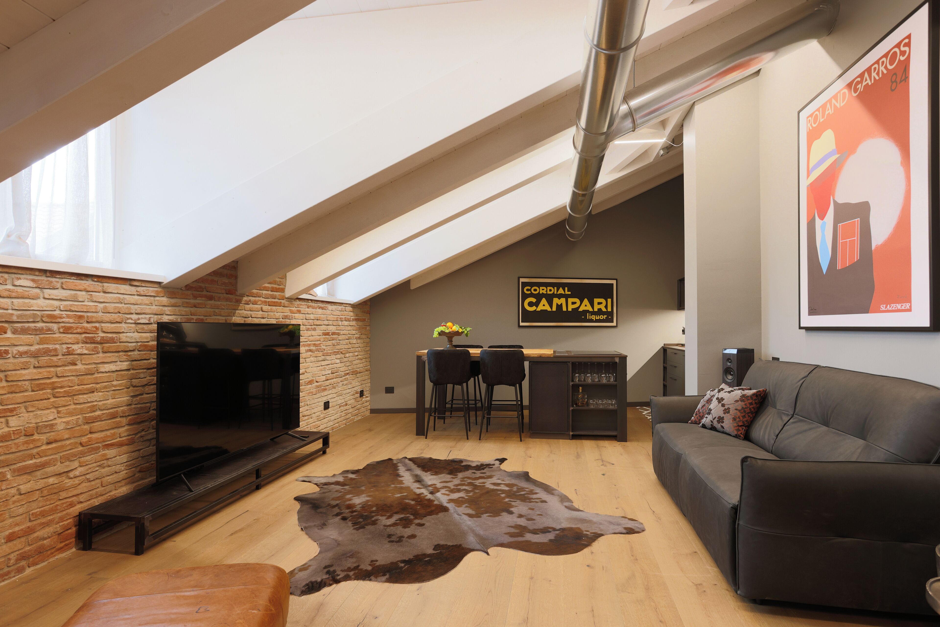 A loft-style living room with a brick wall, a flat-screen TV, a leather sofa, a cowhide rug, and a bar area with stools and a liquor sign.