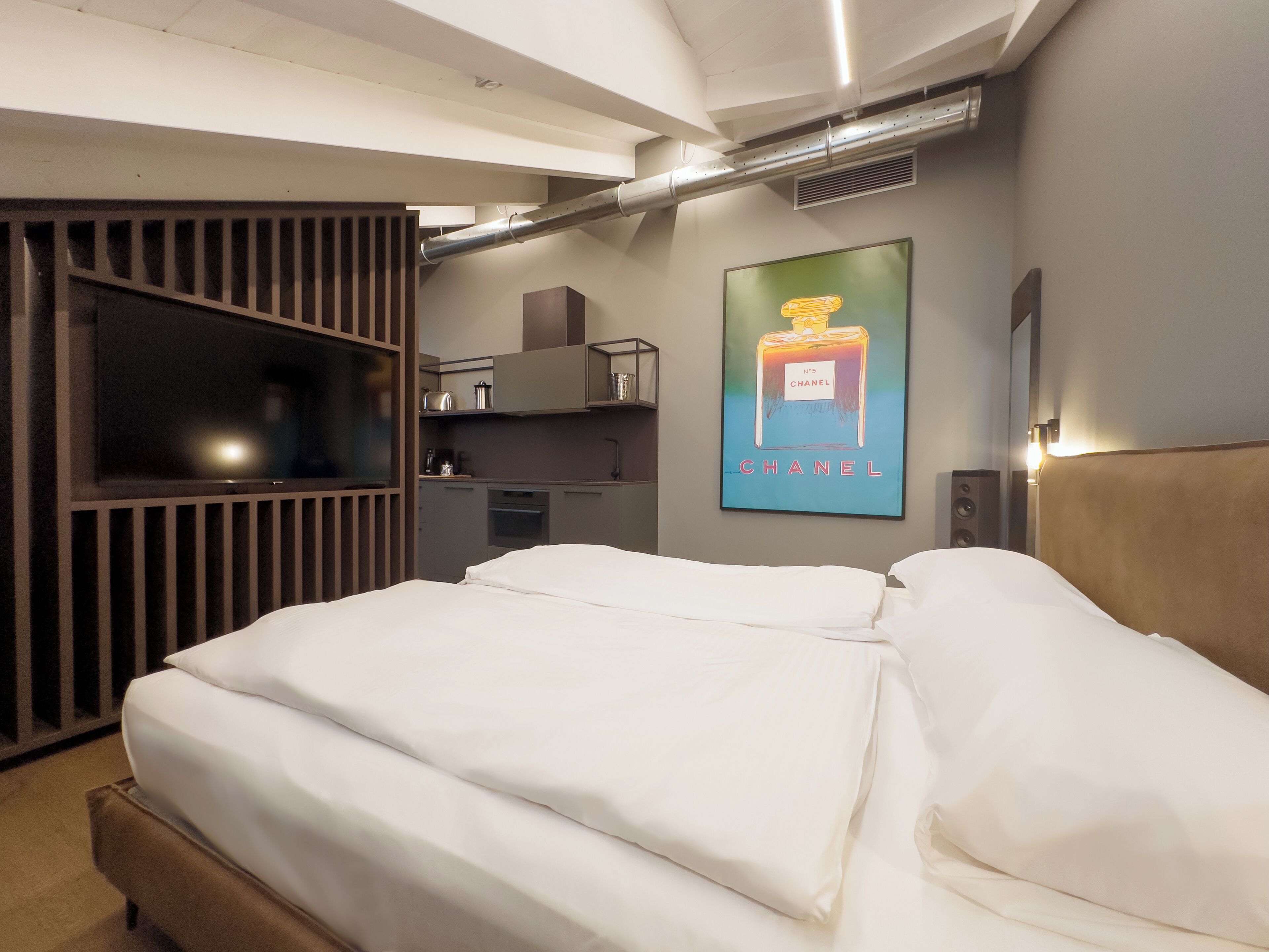 A modern hotel room with a bed, a TV, a kitchenette, and a poster of Chanel perfume.
