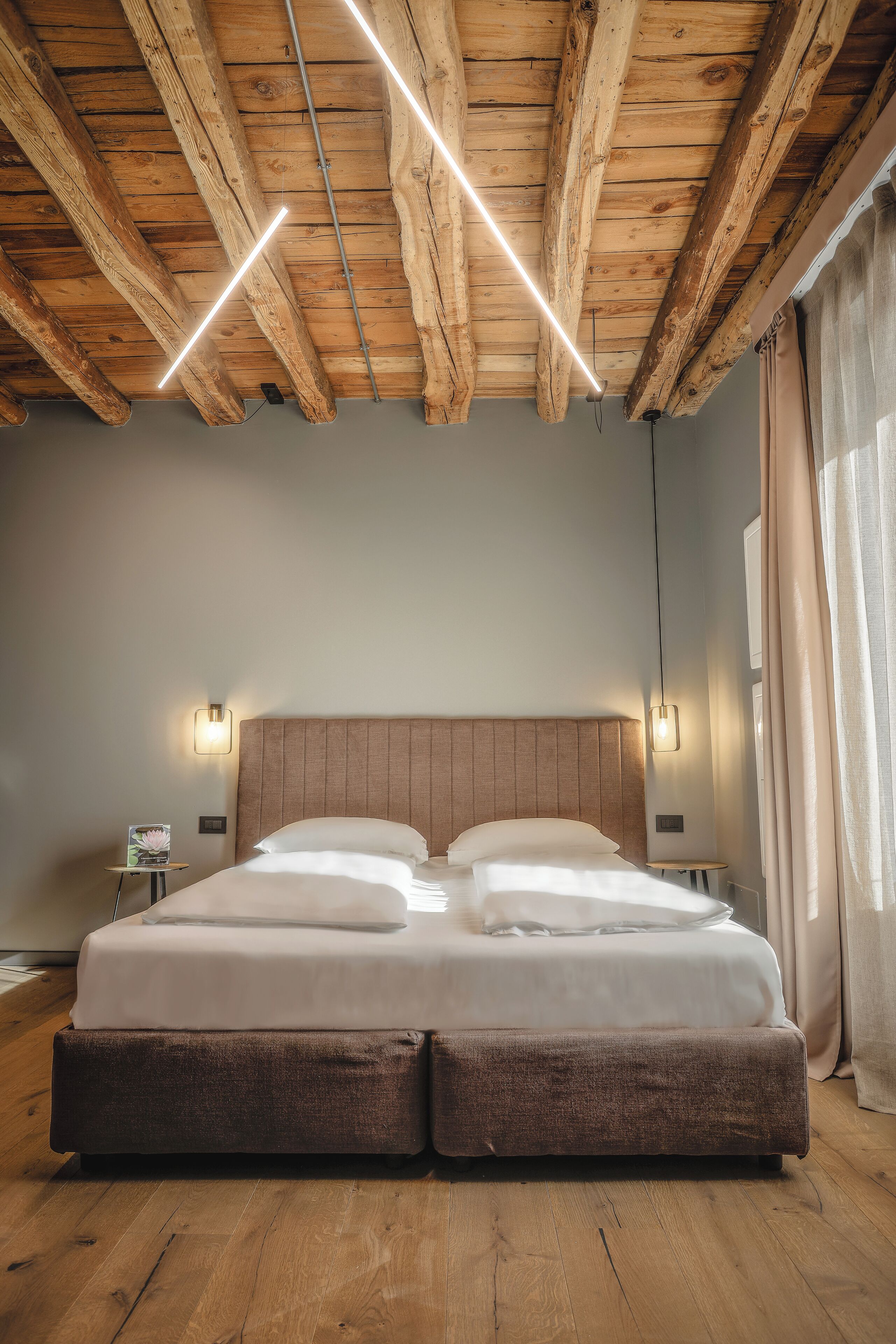 Un dormitorio con techo de madera, una cama grande con cabecera, mesitas de noche y una ventana con cortinas.