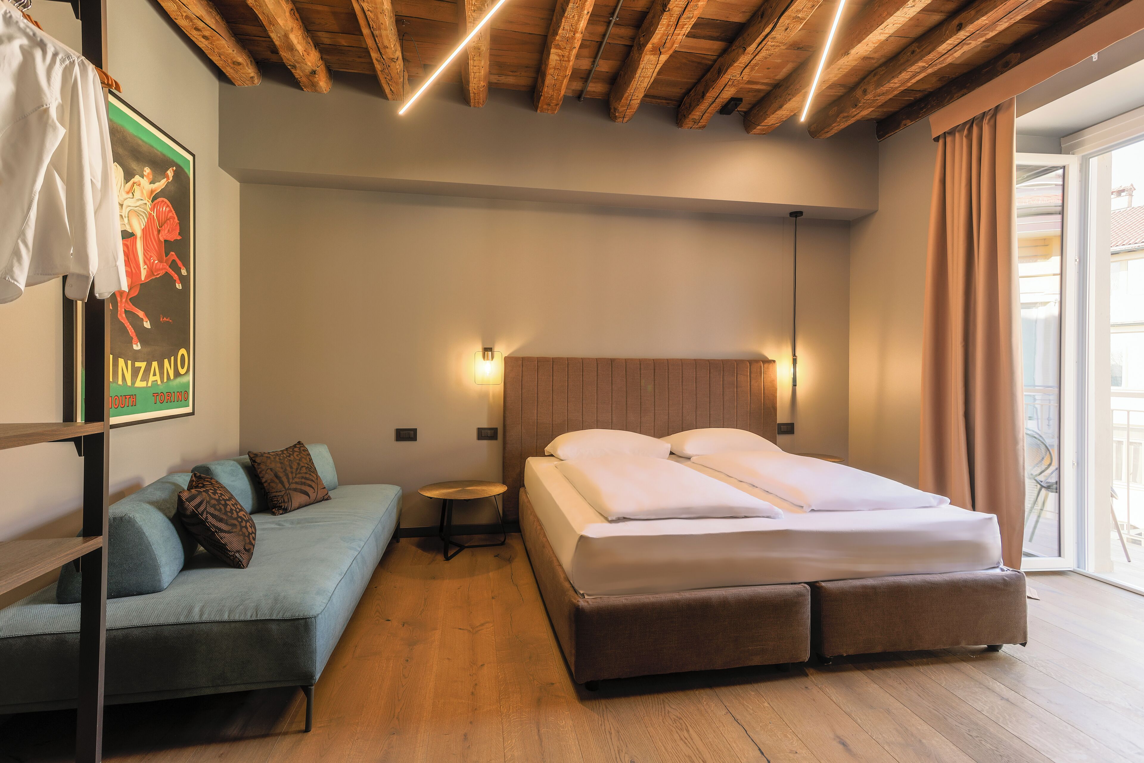 Quarto de hotel moderno com teto de madeira, cama com roupa de cama branca, sofá azul com almofadas, mesa redonda pequena e um cartaz na parede.