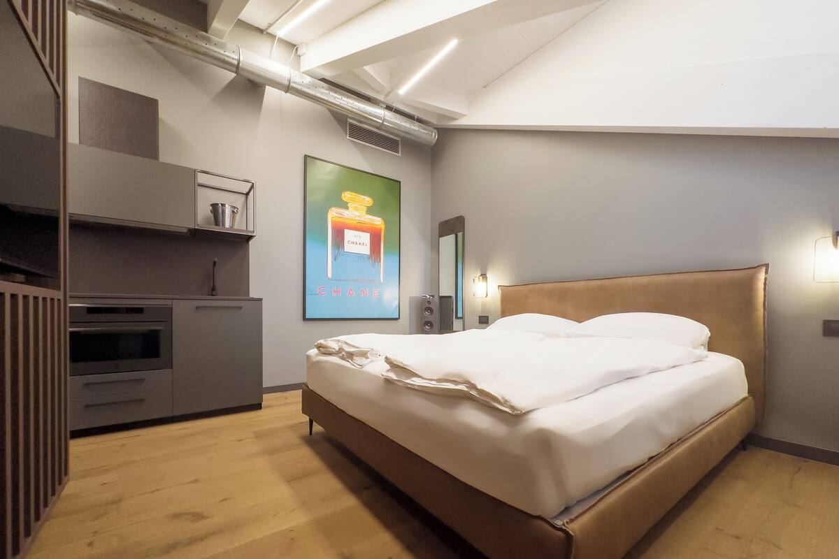 Moderni hotellihuone, jossa on puulattia, valkoisilla lakanoilla varustettu sänky, sisäänrakennettu vaatekaappi, pieni keittiö ja suuri juliste, jossa on kuva hajuvesipullosta.