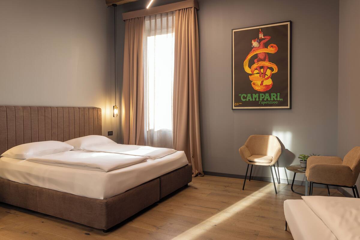 Hotellihuoneessa on sänky, kaksi tuolia, pieni pöytä, jossa on kasvi, ja seinällä juliste.