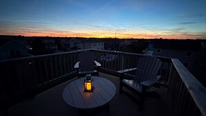 En terrass med två Adirondack-stolar och ett runt bord, med utsikt över en solnedgång över ett bostadsområde.