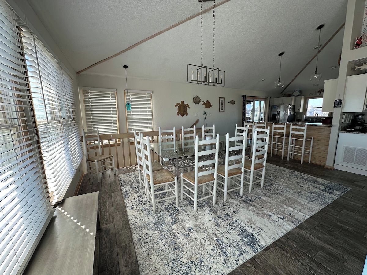 A spacious dining area with a glass table surrounded by white chairs, a patterned rug, and a kitchen in the background.